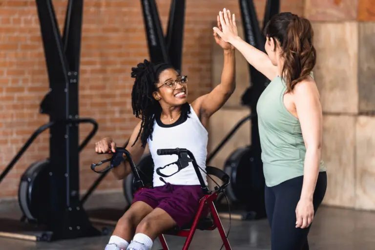 A woman in a wheelchair high-fives their personal trainer after exercising together in a gym